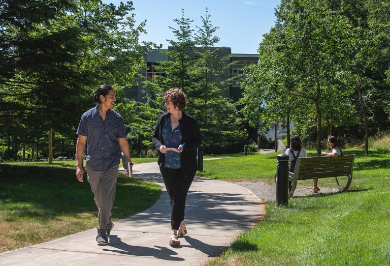 Two people share a path at Royal Roads University, surrounded by green grass and pine trees.