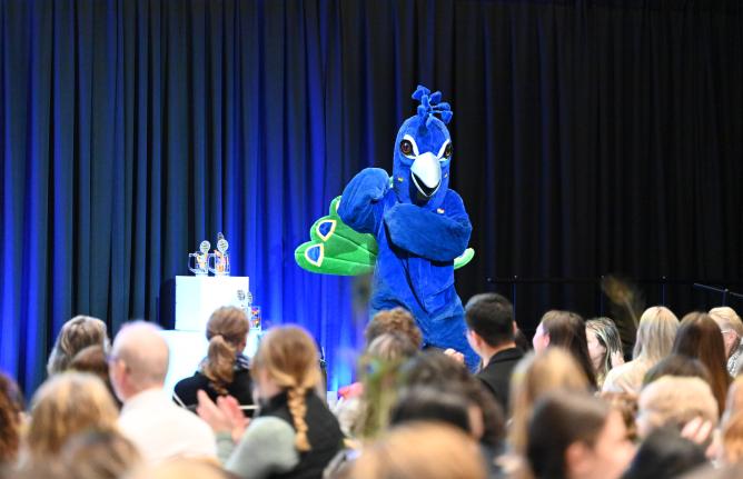 Peacock mascot on a stage in front of a crowd of people