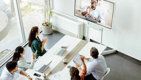 a group of people sitting around a conference table looking at a colleague working virtually through Zoom